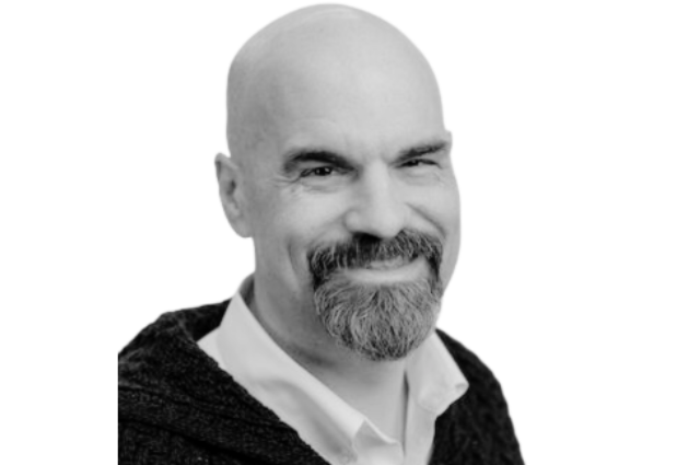 Micheal smiling warmly, bald with a goatee, wearing a dark sweater over a white shirt