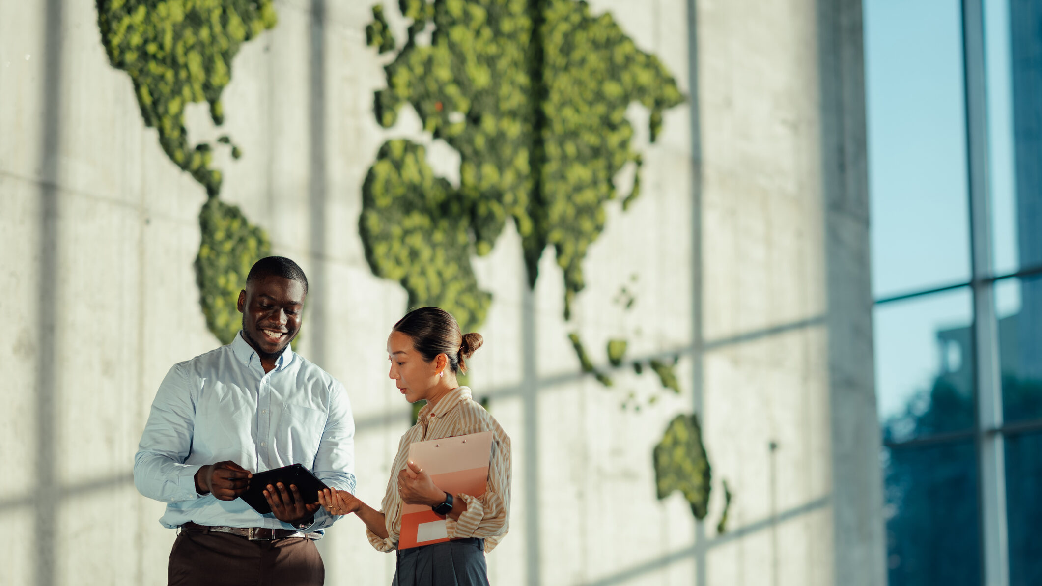 Two colleagues discussing a tablet in front of a green world map wall