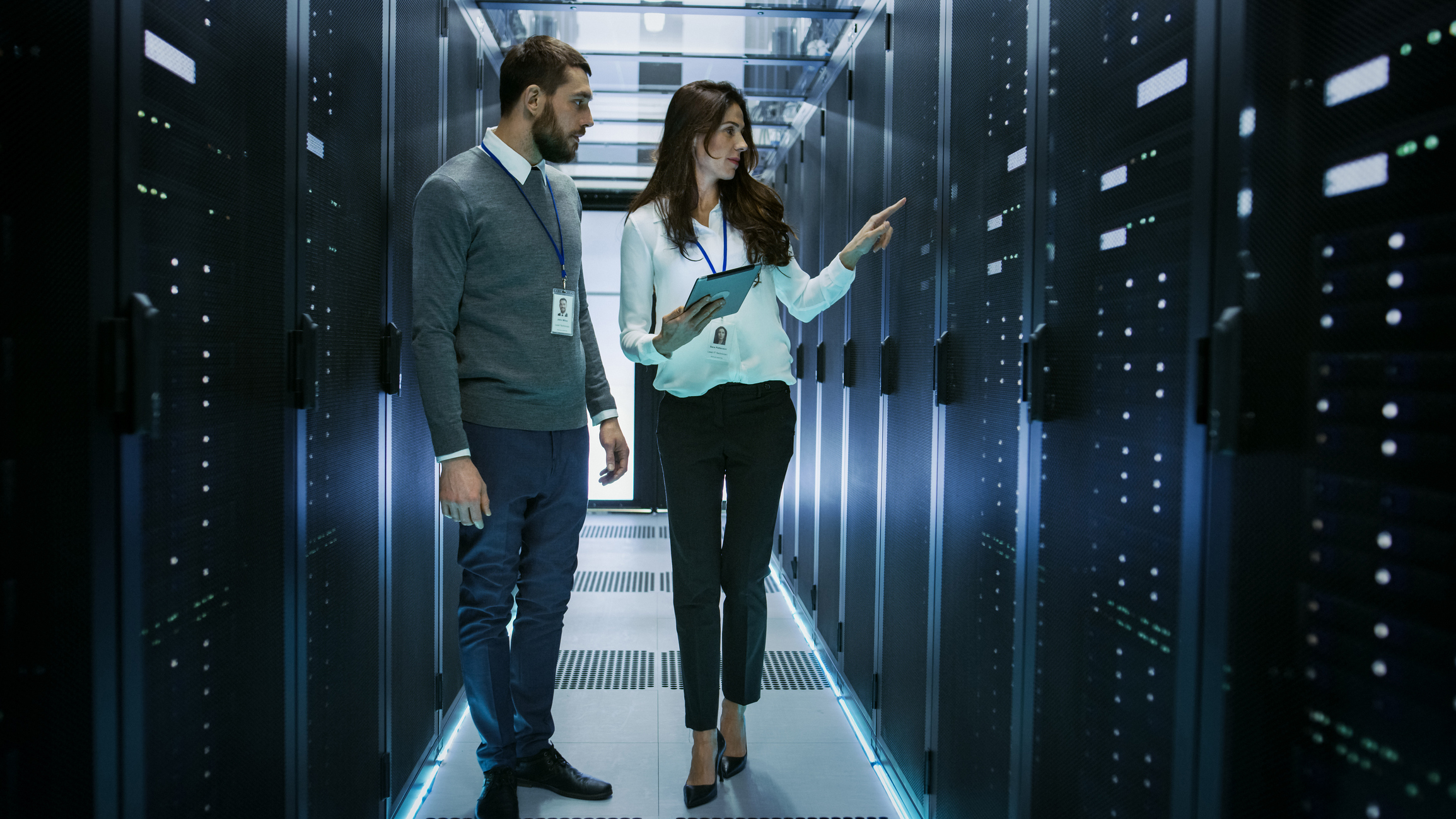 Two people in a server room, checking equipment, holding tablet and wearing ID badges