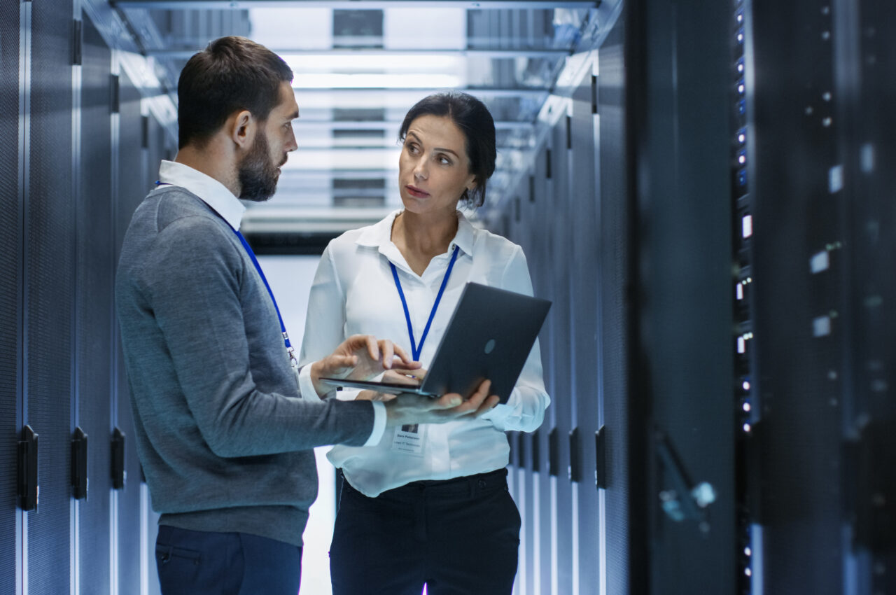 Two people discussing in a data center hallway, one holding a laptop