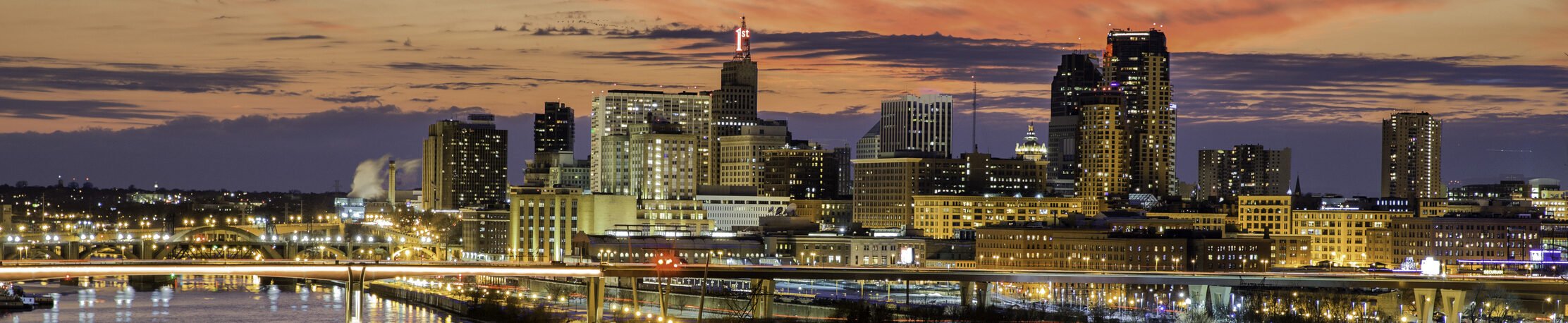 Sunset view of a city skyline with illuminated buildings and a brightly lit bridge crossing a river